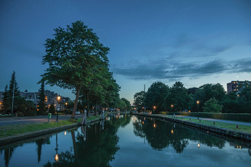 Obraz premium Night view of wide tree-lined canal, buildings and lamp post lighting at dawn in Weesp. Quiet and pleasant village full of canals and green near Amsterdam. Northern Netherlands.