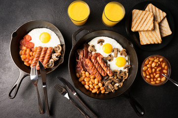 Full English breakfast for two persons: fried eggs with bacon, mushrooms, beans, grilled sausages, toast and orange juice on a black stone background. Flat lay. Top view.
