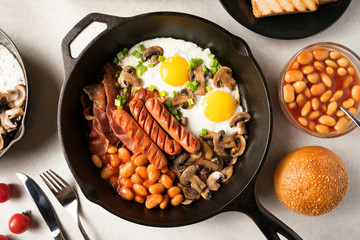 Full English breakfast with fried eggs, sausages, bacon, mushrooms, beans in cast-iron frying pan with toasts and orange juice on white stone background. Close up. Flat lay. Top view.