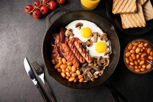 Full English breakfast with fried eggs, sausages, bacon, mushrooms, beans in cast-iron frying pan with toasts and orange juice on black stone background. Close up. Flat lay. Top view.
