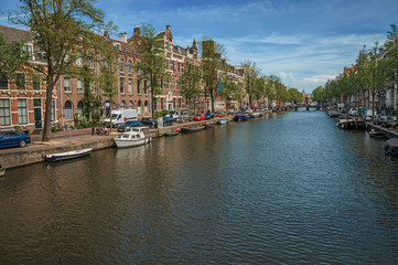 Fototapeta premium Tree-lined canal with old brick buildings, bridge, moored boats and sunny blue sky in Amsterdam. The city is famous for its huge cultural activity, graceful canals and bridges. Northern Netherlands.