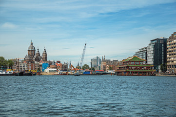 Port with ships, church towers and pagoda building restaurant on canal under a sunny blue sky in Amsterdam. Famous for its huge cultural activity, graceful canals and bridges. Northern Netherlands.