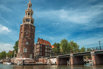 Old brick bell tower and bridge near the tree-lined canal with moored boats and blue sky in Amsterdam. Famous for its huge cultural activity, graceful canals and bridges. Northern Netherlands.
