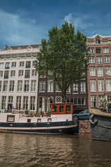 Boats moored at side of tree-lined canal, old buildings and sunny blue sky in Amsterdam. The city is famous for its huge cultural activity, graceful canals and bridges. Northern Netherlands.