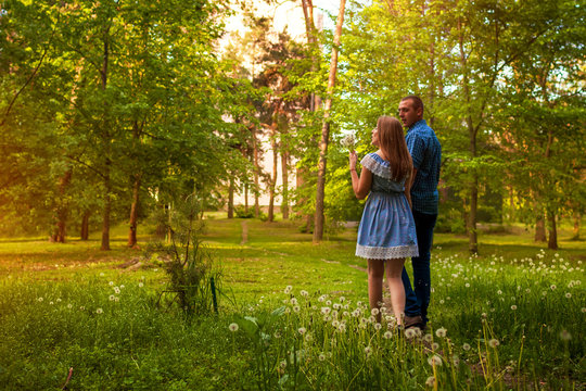 Couple walking in spring forest. Young man and woman blowing dandelions at sunset.