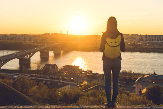 Woman Tourist With A Backpack Look On The Sunset From The High, And Enjoy It In A Golden Hour In Nizhny Novgorod City. View On City And Bridge Across The Oka River. Full-length Rear View On A Person