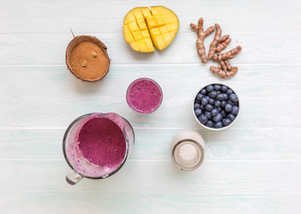 Obraz na płótnie Canvas Mango coconut blueberry turmeric smoothie in dlass bowl with ingredients, coconut milk in glass bottle, coconut sugar in half coconut shells, berries, turmeric root on light blue table, 