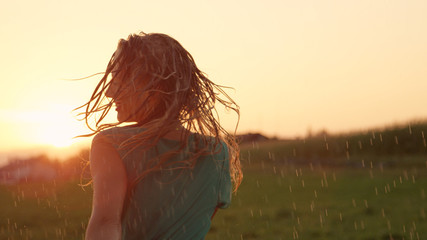 CLOSE UP: Carefree blonde woman dancing outdoors in the fresh rain at sunset.