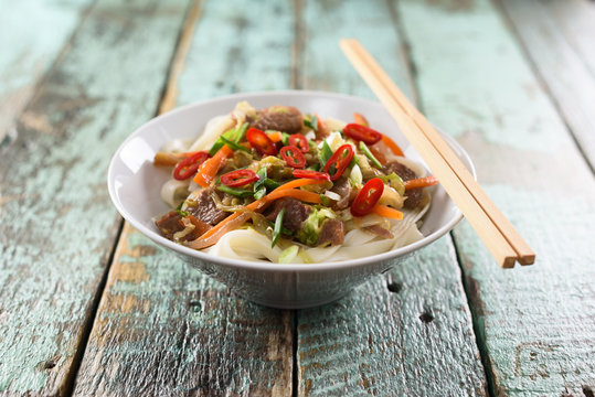 Traditional Asian noodles. Udon stir fry noodles with meat, vegetables and chili in bowl with chopsticks on rustic wooden background