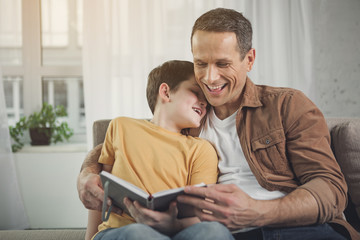 Joyful parent is embracing boy while holding a book. They are sitting on couch and smiling