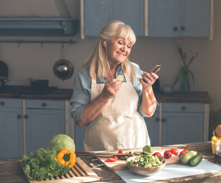 Portrait of happy senior lady listening to music from headphones while cooking in kitchen. She is looking at mobile phone and smiling while eating cucumber slice