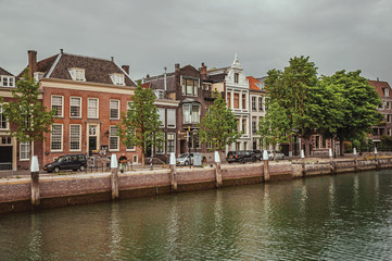 Fototapeta premium Wide tree-lined canal with brick buildings in a street next to it and cars in a cloudy day at Dordrecht. Important and historic port city bordered by rivers. Southern Netherlands.