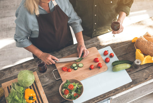 Top view of cheerful mature couple standing in kitchen. Woman is cutting vegetables while man is holding a wineglass. Focus on healthy food on table