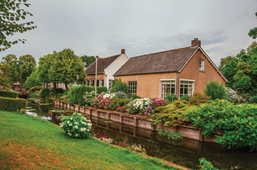 Charming small canal next to rustic houses with lush flowery garden and lawn in cloudy day at Drimmelen. A lovely small hamlet with harbor and elegance streets. Southern Netherlands. 