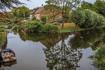 Obraz premium Charming small canal next to rustic houses with lush gardens and trees reflected on the water in cloudy day at Drimmelen. A lovely small hamlet with harbor and elegance streets. Southern Netherlands. 