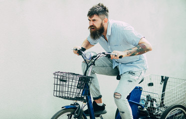 Bearded man in denim outfit on white wall. Delivery guy trying hard to be on time. Hipster with tattooed arm riding bike, urban scene. Man in torn blue jeans cycling on street, active leisure concept
