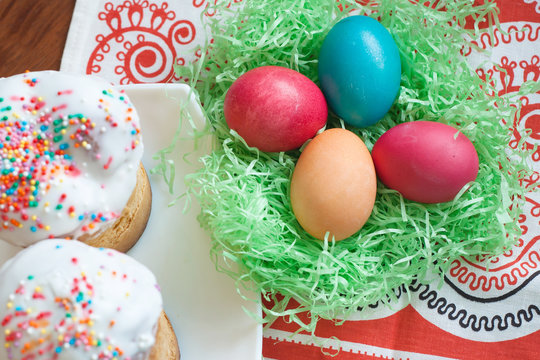 Top view of Easter cakes, next to colorful colored Easter eggs. Painted chicken eggs are in artificial decorative straw. Festive symbols on the dining table.