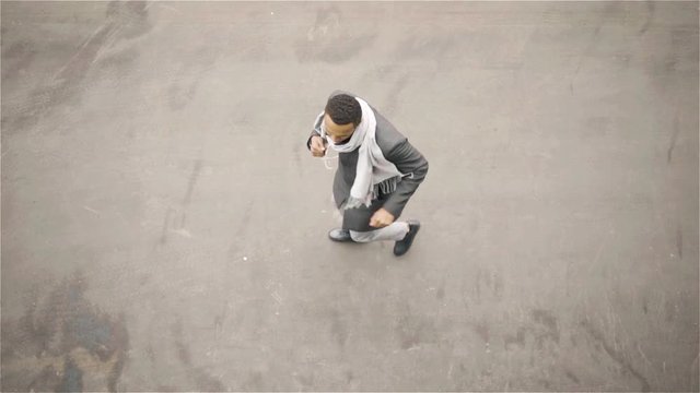 Top view of a cheerful young African American man wearing a coat and a scarf dancing in an autumn street. He is listening to the music in his headphones. Handheld top view shot 60FPS