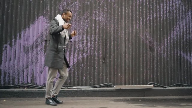 Cheerful young African American man wearing a coat and a scarf dancing in an autumn street. Handheld medium shot 60FPS