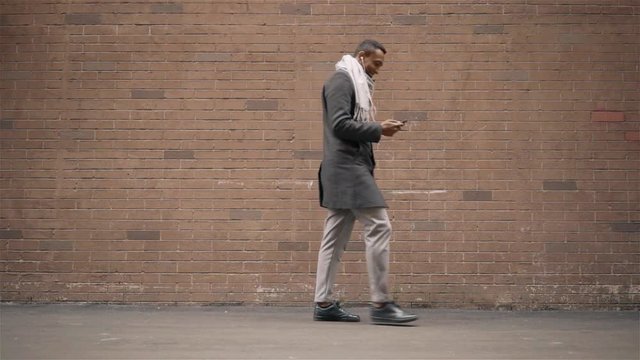 African American man in a coat and scarf listening to music and walking along a brick wall on an autumn day. Tracking slow motion medium shot