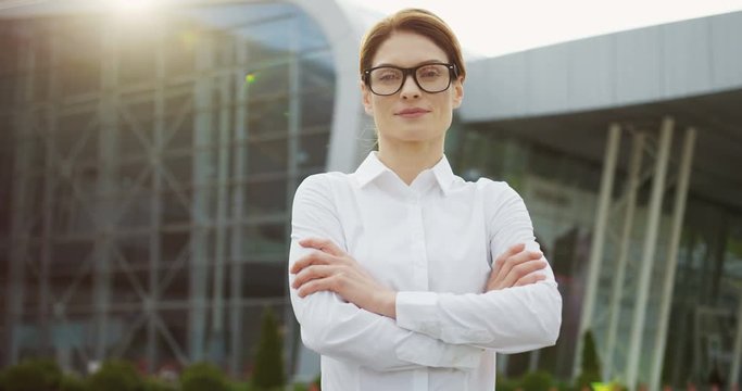 Portrait shot of the Caucasian pretty business lady smiling to the camera and crossing her hands while standing on the huge glass building background. Outside