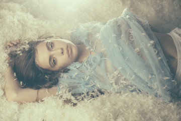 Beautiful girl caressing her skin. Young female in blue silk dress lying on soft pile of white feathers, bedtime fantasy. Tenderness and intimacy concept