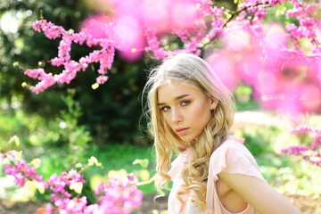 Fototapeta premium Beautiful girl standing under gentle tree with tiny hot pink blossom. Charming blond young woman breathing fresh aroma of blooming flowers outdoors, spring time concept