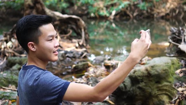 Selfie photo concept : Asian young man holding smartphone for selfies at  forest, beautiful scenery for peaceful in spring , It shows moisture, tranquility and refreshing of rain forest in Thailand