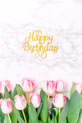 Pink tulips and gold Happy birthday letters on white marble background. Spring and celebration concept. Copy space