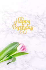 Pink tulips and gold Happy birthday letters on white marble background. Spring and celebration concept. Copy space