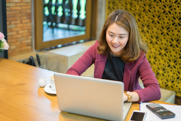 Smiling beautiful Asian girl with black hair working in cafe,Casual woman manager sitting in her office workspace with computer and big bright window and drinking coffee ,look at laptop,vintage color