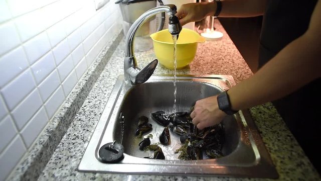 Male hands taking off byssus from mussels under the running water in sink. Seafood preparation process.