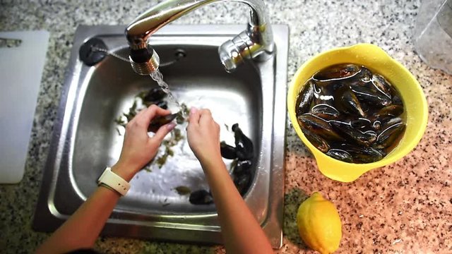 Top view of woman hands removing weed from mussels under the running water in sink and then putting them into bowl with water. Seafood preparation process.