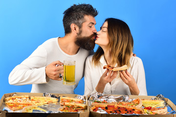 Obraz na płótnie Canvas Loving couple sitting at table kissing and eating pizza. Pretty young girl in white shirt holds slice yummy pizza and handsome boyfriend in white pullover with glass of beer. Boxes with pizza on table
