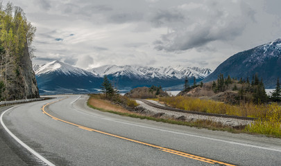 Alaska Scenic Road:  The Seward Highway curves beneath cloudy skies as it passes by snow-covered mountains at the edge of an ocean inlet south of Anchorage.