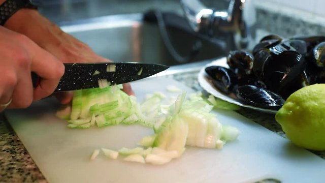 Close-up of young chef hands with knife chopping fresh onion into small cubes on a white cutting board with mussels on background.