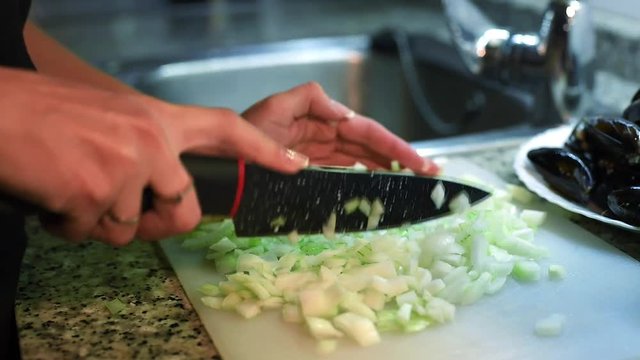 Slow motion of young chef hands with knife chopping fresh onion into small cubes on a white cutting board with mussels and lemon on background.