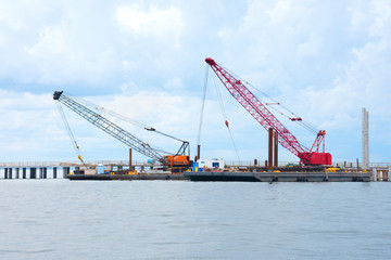 Bridge construction being done with two building crawler cranes on flat top boats floating on calm water in a bay or river.