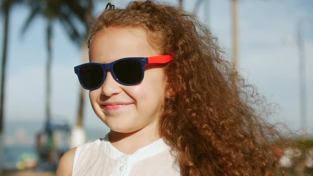 Close-up portrait of a happy cute little girl child with curly hair and red sunglasses looking into the camera