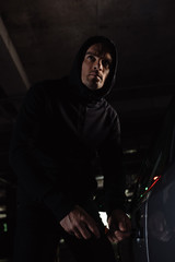Obraz na płótnie Canvas cautious male robber in black hoodie intruding car by screwdriver