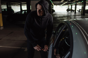 cautious male robber in black hoodie intruding car by screwdriver