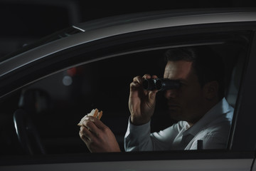 male private detective doing surveillance by binoculars and eating sandwich in car
