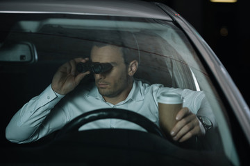 undercover male agent doing surveillance by binoculars and drinking coffee in car