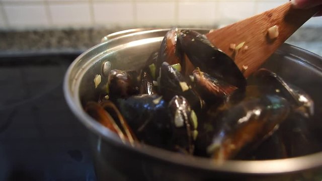 Close-up of female hands stirring mussels with wooden spatula in the pan with white sauce of fried onion, garlic and wine. Seafood cooking.