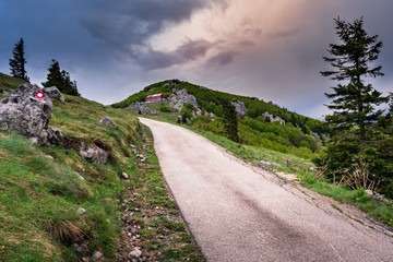 View of a road leading uphil to a mountain or mountaineer's hut on the Velebit mountain in Croatia during sunset on a rainy cloudy day, mountain trail sign. Travel and nature concept