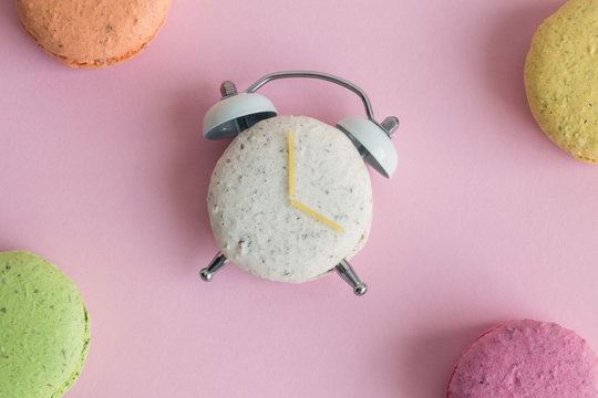 Flat lay of colorful macarons on pastel pink background. One of them in shape of alarm clock with clock hands made of spaghetti.