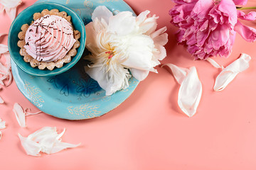 Picturesque still life in pink tones, decorative plate with creamy cake surrounded by white petals pions, nearby is a pink flower, on a pink background, copy space, top view, set