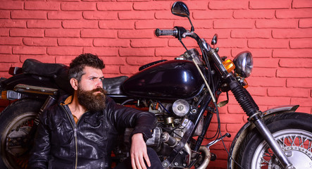 Fototapeta na wymiar Man with beard, biker in leather jacket near motor bike in garage, brick wall background. Bikers lifestyle concept. Hipster, brutal biker on pensive face in leather jacket sits, leans on motorcycle.