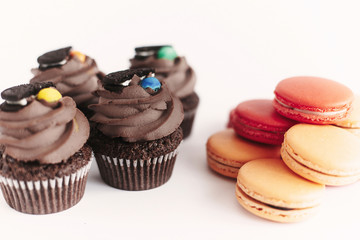 stylish chocolate cupcakes with cookie and candy on top and colorful macaroons on white background. space for text. catering for celebration concept