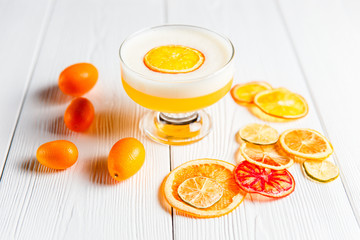 A photo of a bright orange alcoholic drink in a cocktail glass, garnished with dried orange, lime and grapefruit and fresh cumquat. White wooden table.